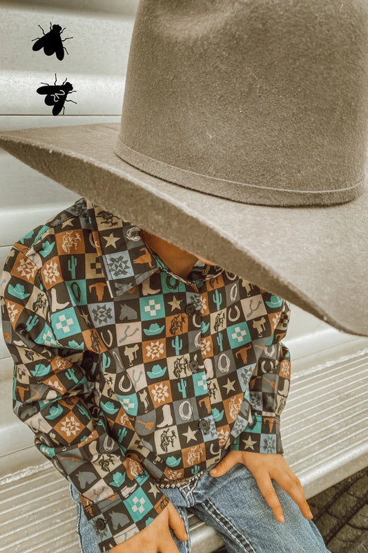 A boy wearing a cowboy hat is leaned into the camera with his face out of frame. His button-up shirt is checkered with different images such as cacti, saddles, cowboy boots, and lasso rope. He is sitting on a trailer.