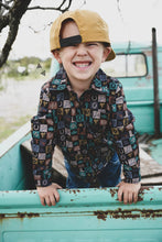 Load image into Gallery viewer, A boy is in the bed of a vintage pick-up truck. He has a giant grin on his face, with an oversized baseball cap backward on his head. The shirt is checkered with various images such as saddles, lasso rope, horses, cacti, and other western icons.