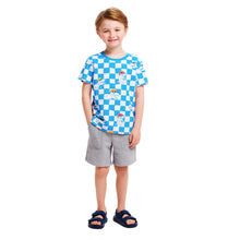 Load image into Gallery viewer, A young boy wearing A cotton crewneck t-shirt with a red neck hem on the inside of the shirt. The main design features featuring blue and white checkers, with several t-rex dinosaurs integrated into the design, with the dinosaurs wearing fitted hats and sunglasses. He is also wearing open-toed sandals, and grey knit shorts. He has his hand in his pocket.