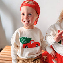 Load image into Gallery viewer, A cheerful toddler wearing a cream sweater with a red truck carrying a Christmas tree design, paired with a backward red cap, sitting on a wooden bench and smiling brightly.