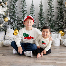Load image into Gallery viewer, Two siblings sitting in front of a festive holiday backdrop. The older brother wears a cream sweater with a red truck and tree design and a matching cap, while the baby sibling wears a similar sweater. Decorated Christmas trees and gold ornaments surround them.