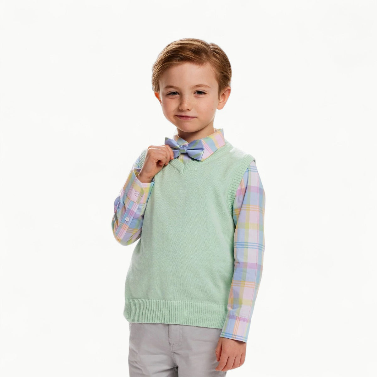 A smirking male child with strawberry blonde hair is wearing a pastel-colored outfit, including a blue and green plaid Bow-tie, a pink plaid button up shirt, and a pastel green sweater vest. He is also wearing light grey chinos.