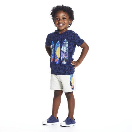 A male child wearing a Navy blue short-sleeved hooded t-shirt with dark navy palm tree pattern. The front features three brightly colored surfboards. He has paired the outfit with white shorts that have the same surfboard pattern. He also has on navy blue sneakers. 