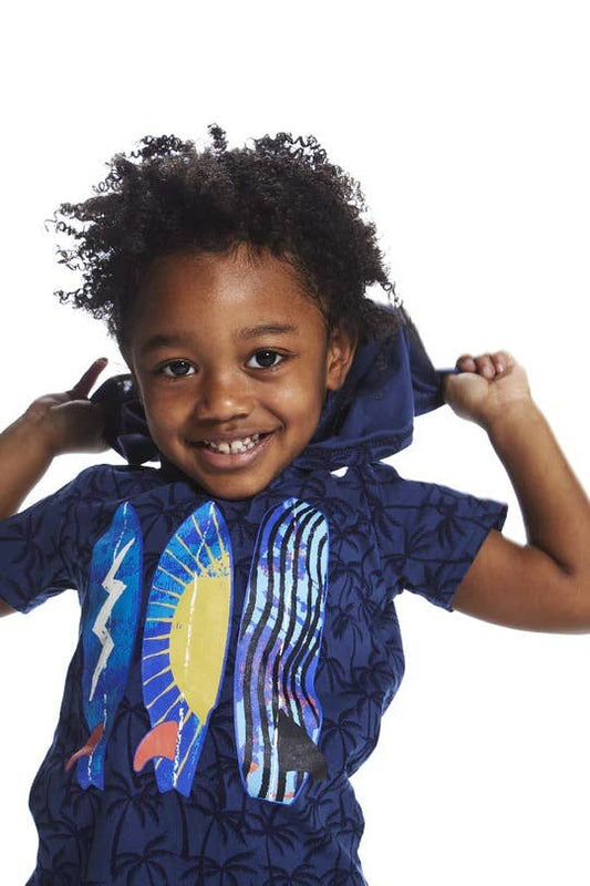 A smiling male child wearing a Navy blue short-sleeved hooded t-shirt with dark navy palm tree pattern. The front features three brightly colored surfboards. 
