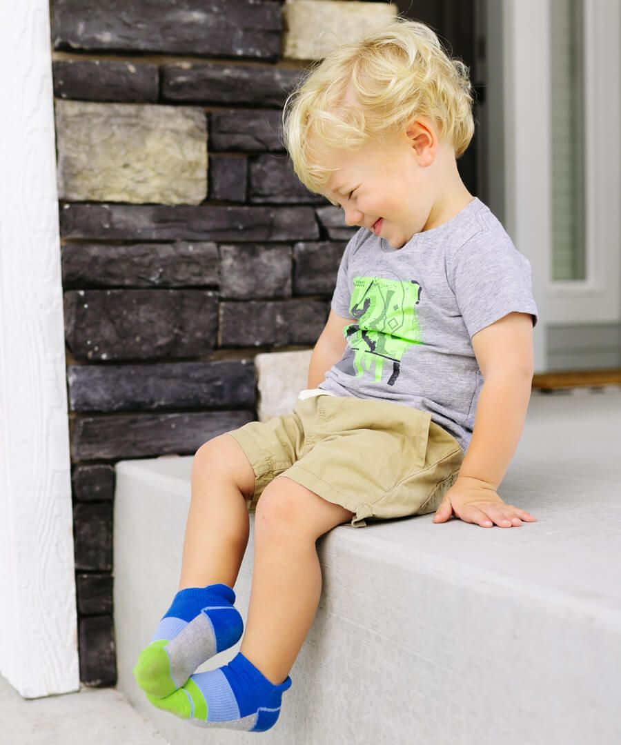 A male toddler sitting on a stoop outside a home. The toddler is blonde, and is grinning. He has on a gray graphic tee, and khaki shorts. The toddler is wearing a blue and green pair of ankle socks.