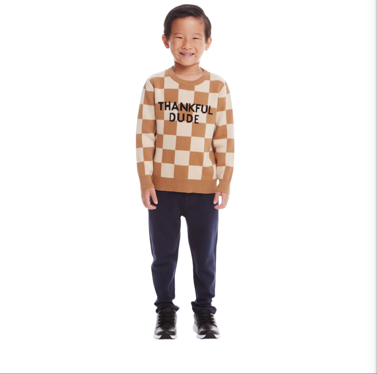 Young boy wearing a tan and beige checkered sweater. Black letters on the front say "Thankful Dude." He is wearing navy pants and black shoes. 