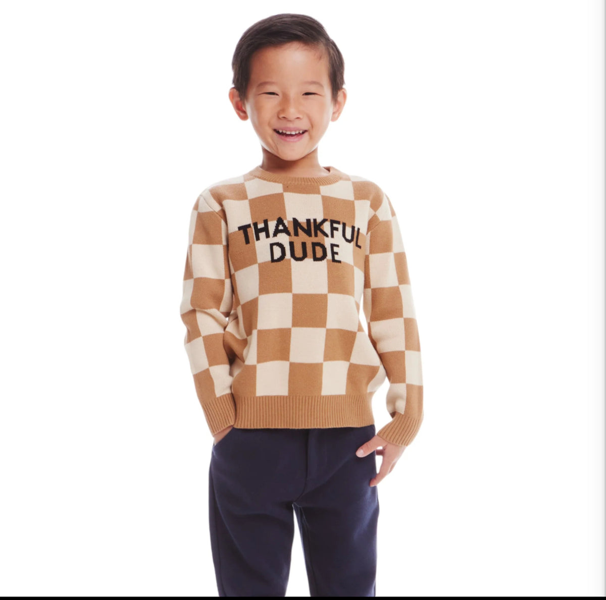 Young boy wearing a tan and beige checkered sweater. Black letters on the front say "Thankful Dude." He is wearing navy pants.