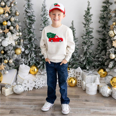 A smiling young boy wearing a festive cream sweater with a red truck and Christmas tree design, paired with a matching red and white cap, jeans, and sneakers, standing in front of a snowy holiday-themed backdrop with decorated Christmas trees and gold ornaments.