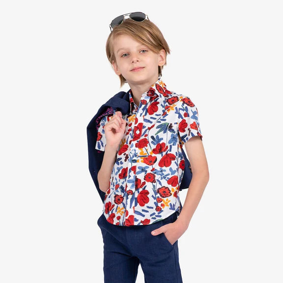 A male child confidently wearing a pair of aviators on his head. His shirt is a white short-sleeve button up shirt adorned with red, yellow, and blue hand painted details. He has a navy blazer thrown across his shoulder, and is also wearing dark navy shorts. 